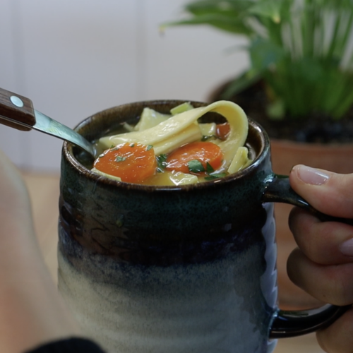 chicken and egg noodle soup in a stone mug with a spoon digging in